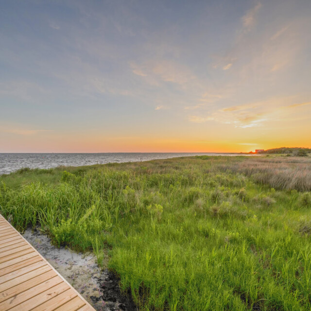 Wooden boardwalk beside grassy marshland at sunset, with calm water and a clear sky in the background.