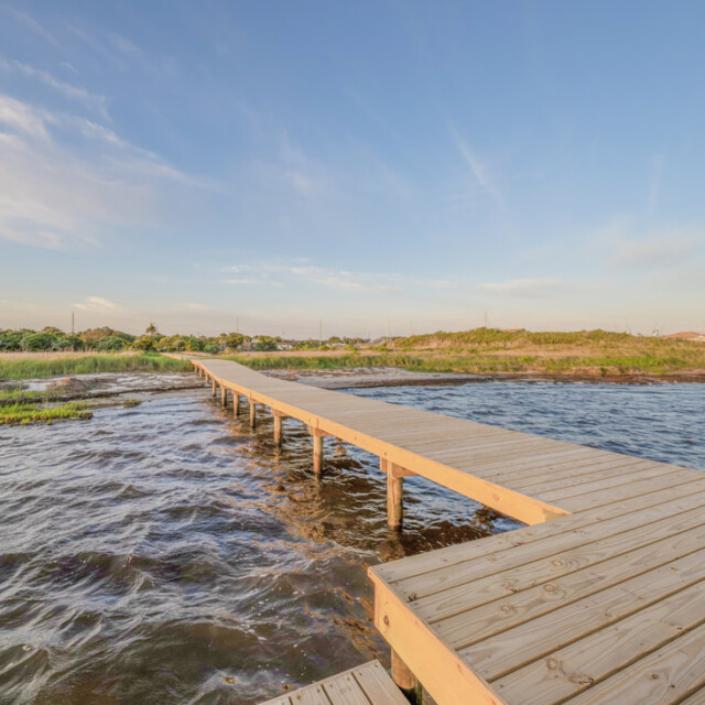 Wooden boardwalk extends over shallow water toward grassy shoreline under a clear blue sky with wispy clouds.