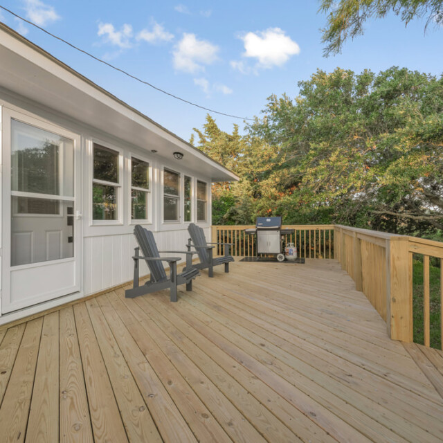 Wooden deck with two black chairs and a grill, attached to a white house, surrounded by trees under a blue sky.