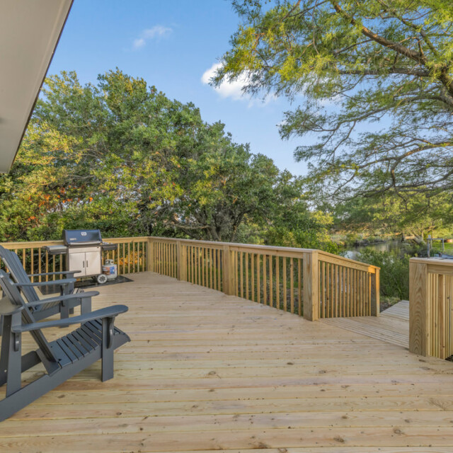 Spacious wooden deck with two chairs, a grill, and a railing, surrounded by trees under a clear sky.
