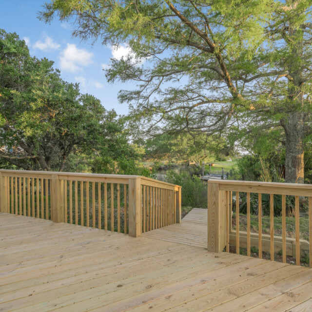 Wooden deck with railing overlooking green trees and a grassy yard on a clear, sunny day.
