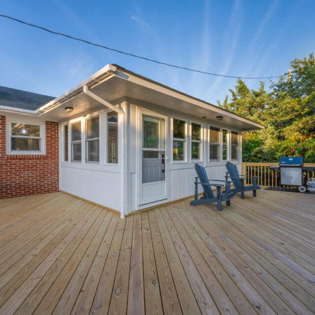 Spacious wooden deck with two chairs, a grill, and a sunroom attached to a brick and white-sided house.