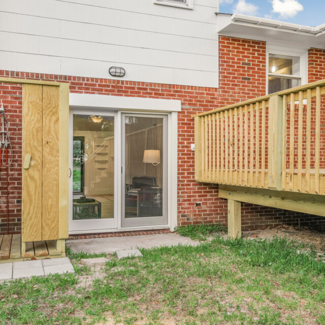 Brick house with sliding glass doors, wooden deck, and outdoor shower area on a small grassy backyard.
