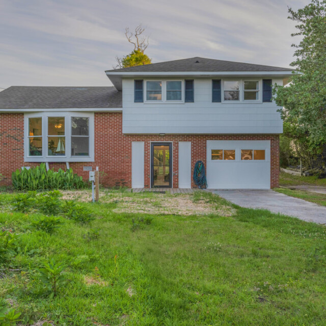 Two-story house with red brick and blue siding, front lawn, driveway, and garage door, surrounded by trees.