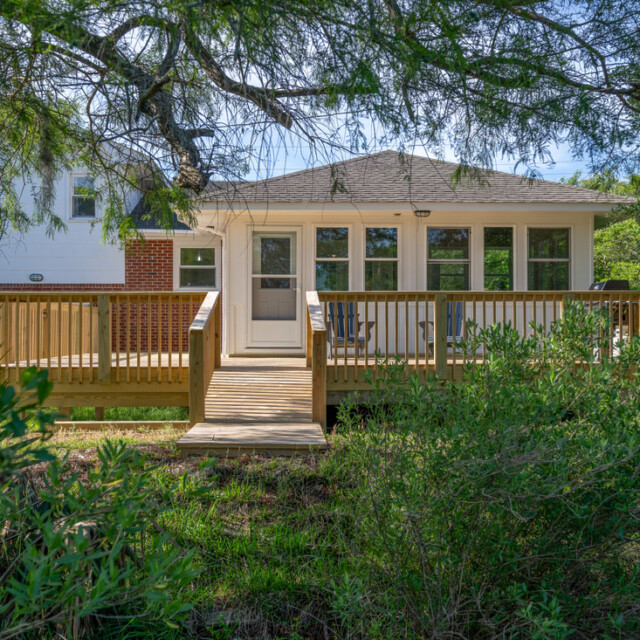 A wooden deck and ramp lead to the back entrance of a house, surrounded by green trees and shrubs.