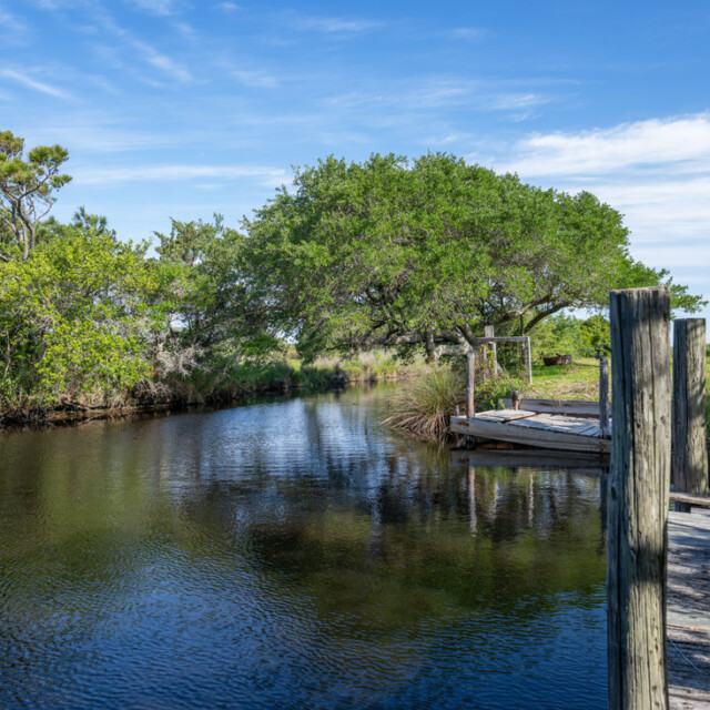 A calm river with a wooden dock, surrounded by green trees under a blue sky with scattered clouds.