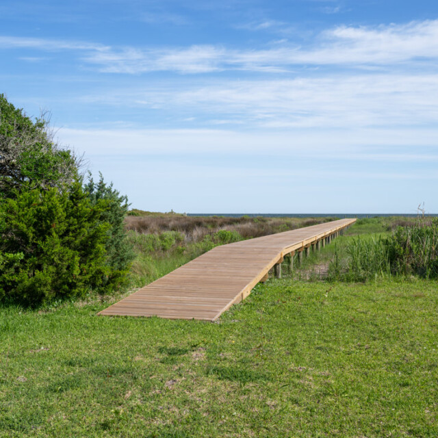 Wooden boardwalk stretching over grassy dunes toward the ocean under a blue sky with scattered clouds.
