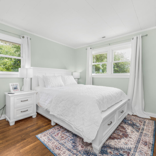 Bright bedroom with a white bed, nightstand, area rug, hardwood floor, and two windows with white curtains.