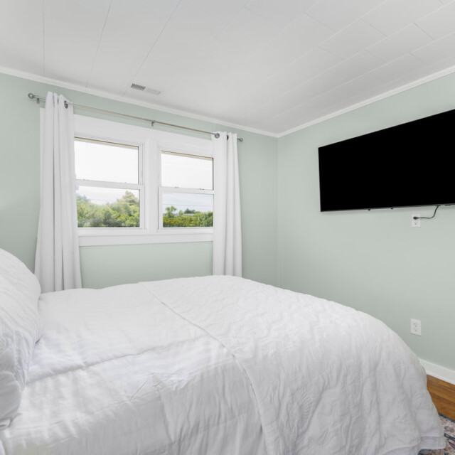 Minimalist bedroom with a white bed, light green walls, two windows, white curtains, and a wall-mounted TV.