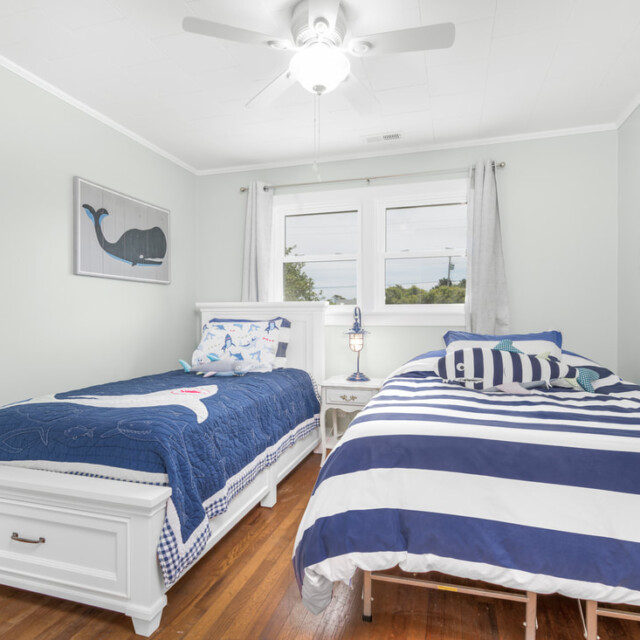 Two twin beds with nautical bedding in a bright bedroom with wood floors, white walls, and a ceiling fan.