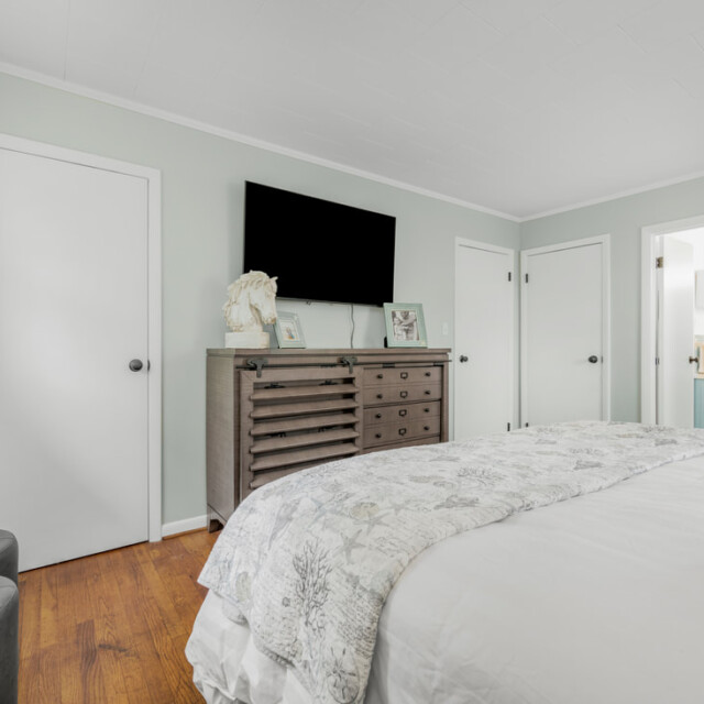 Bedroom with white walls, a bed, a dresser with decor, wall-mounted TV, and two doors leading to other rooms.