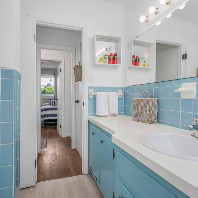 Bathroom with blue tile, white countertop, sink, mirror, and shelves; bedroom visible through doorway.