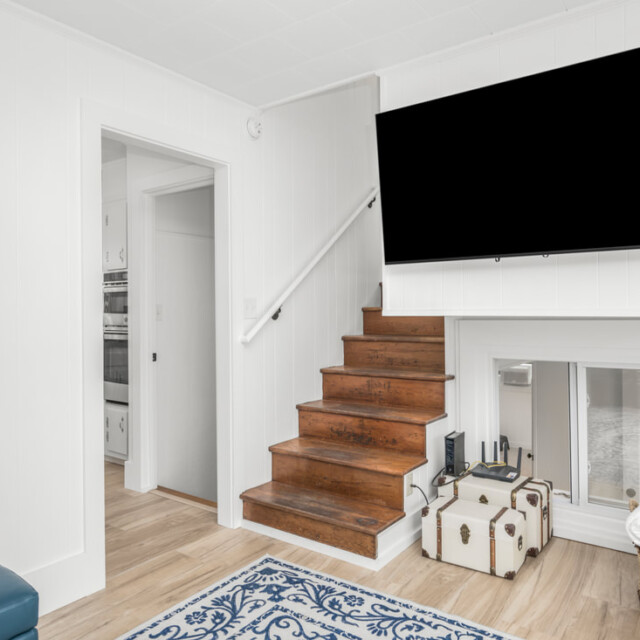 Wood stairs with a mounted TV, decorative trunks, a basket with blankets, and a blue rug in a bright, white room.