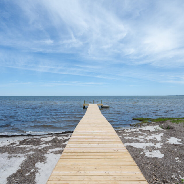 Wooden dock extending over calm blue water with partly cloudy sky and sandy shoreline in the foreground.