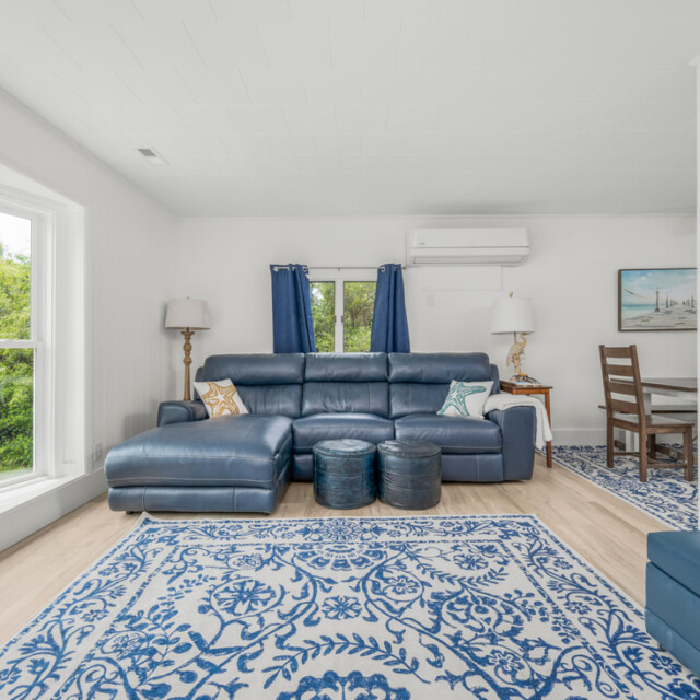 Living room with blue sectional sofa, patterned rug, two ottomans, large window, and a small dining area in back.