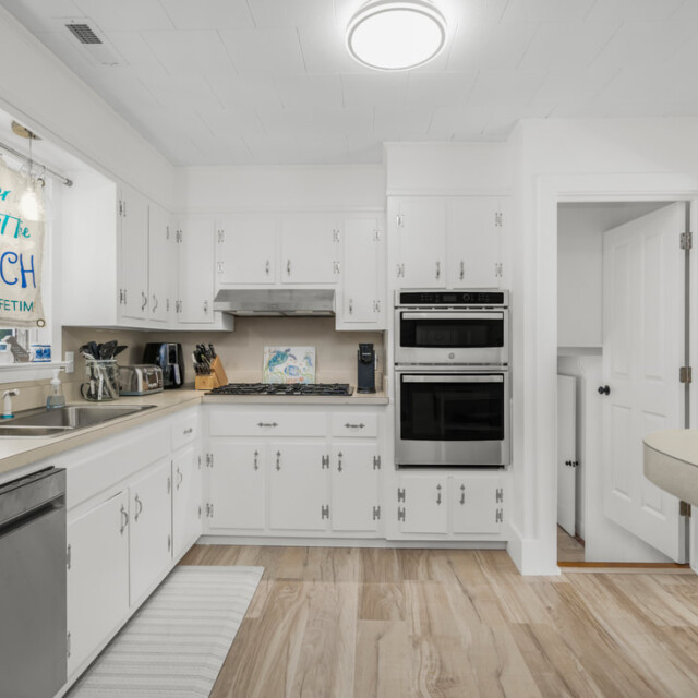 White kitchen with stainless steel appliances, light wood flooring, and a window with a beach-themed sign above the sink.