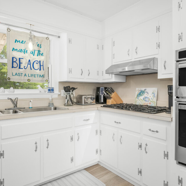 Bright white kitchen with stainless steel appliances, gas stove, double sink, and a “BEACH” sign hanging by the window.