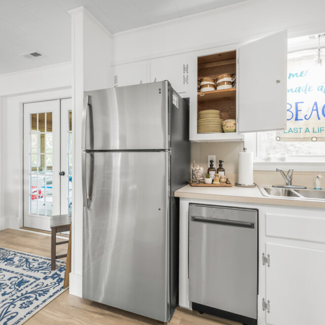 Modern kitchen with stainless steel appliances, open cabinet, and beach-themed decor near a dining area with wooden chairs.