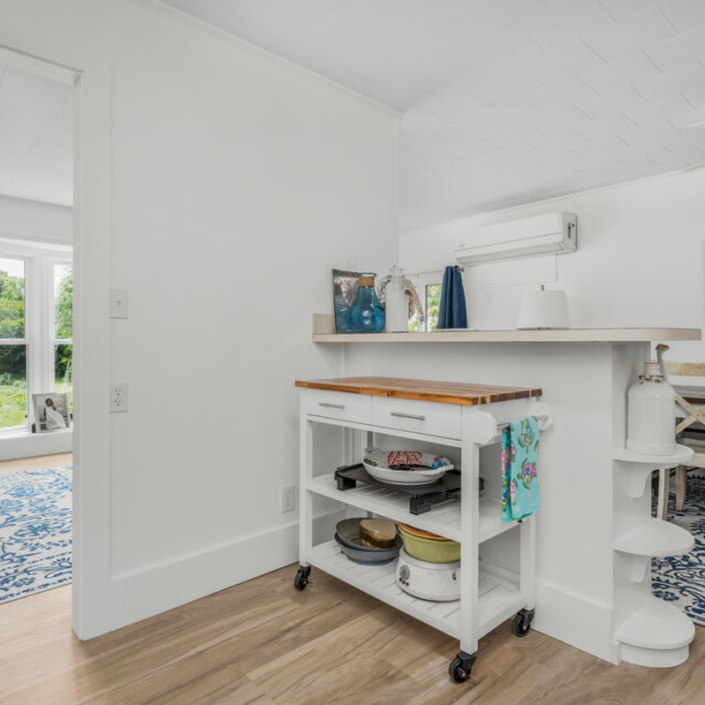 Bright kitchen with a rolling cart, view into dining area, large windows, and blue-patterned rugs throughout.