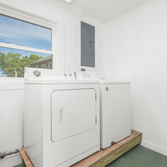 White washer and dryer on a wooden platform in a bright laundry room with a window and electrical panel.