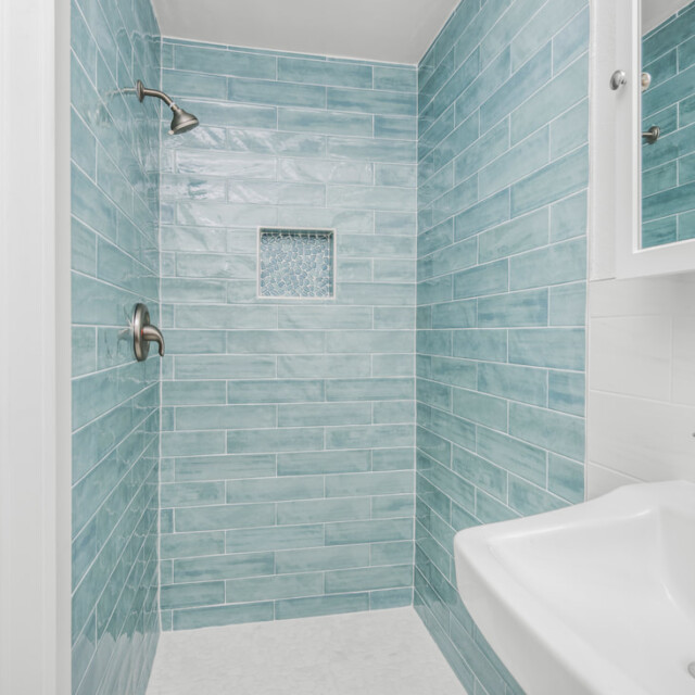 Walk-in shower with blue subway tiles, a built-in niche, and a white sink with a chrome faucet in the foreground.