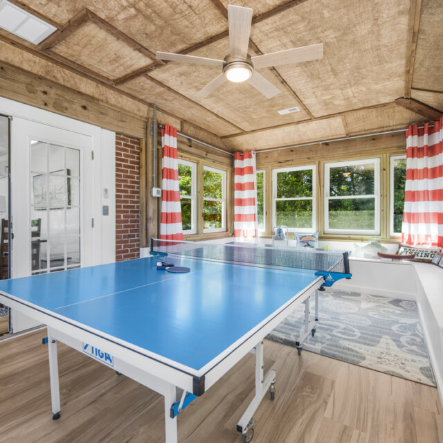 A blue ping pong table in a sunroom with striped curtains, wood accents, and large windows overlooking greenery.