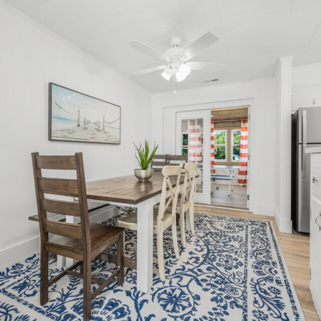 Dining area with wooden table, mixed chairs, blue patterned rug, and view into kitchen with stainless steel appliances.