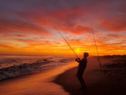 Person fishing on a beach at sunset with vibrant orange and purple clouds in the sky.