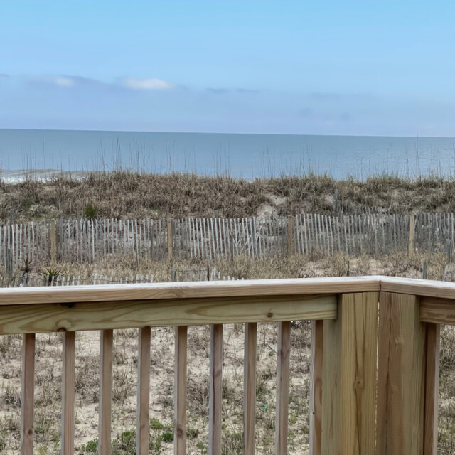 Wooden deck overlooking sandy dunes, weathered fencing, and a calm ocean under a clear blue sky.