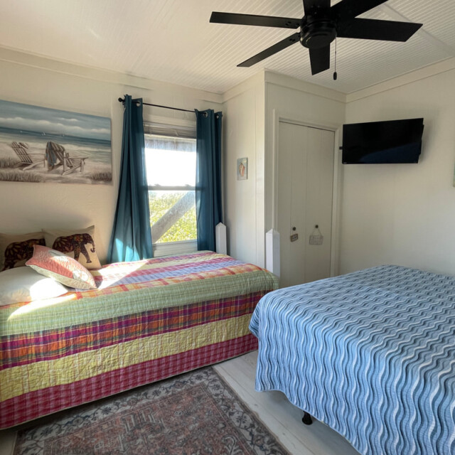 Bedroom with two beds, a window with blue curtains, a ceiling fan, wall art, and a mounted TV.