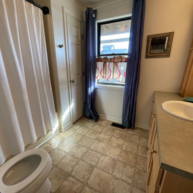 Bathroom with a toilet, sink, shower curtain, window with blue curtains, and beige tile floor.
