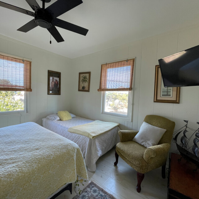 Small bedroom with two beds, armchair, wall-mounted TV, ship model, and three windows with bamboo shades.