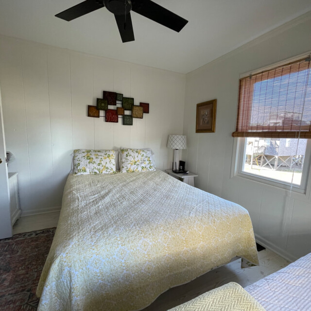 Bedroom with a queen bed, side table, window with bamboo shade, wall art, and ceiling fan.
