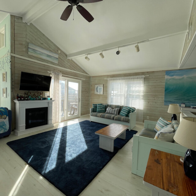 Bright living room with a blue rug, white furniture, TV, fireplace, beach decor, and large windows letting in sunlight.