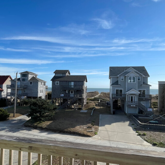 Row of coastal houses on stilts facing the ocean under a blue sky, viewed from a wooden deck railing.