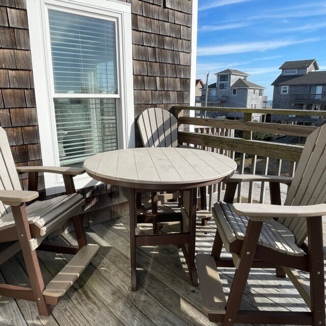 Three wooden chairs and a round table on a balcony with shingle siding, railing, and coastal houses in the background.