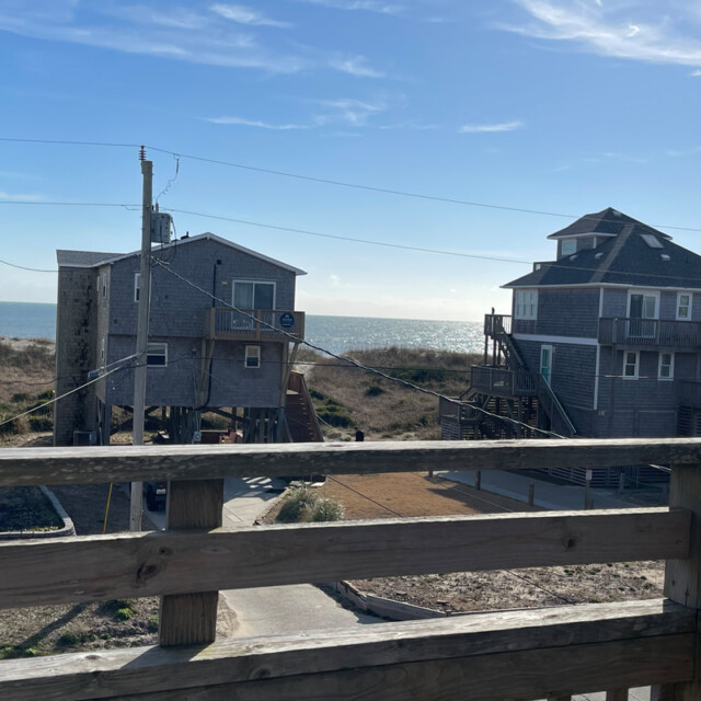 View from a wooden deck overlooking beach houses on stilts with the ocean and blue sky in the background.
