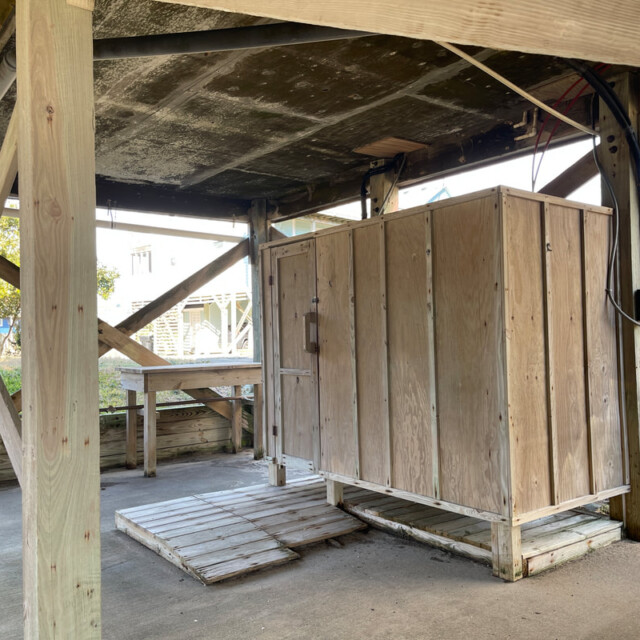 Outdoor wooden shower enclosure and bench under a raised house, on a concrete slab with open sides.