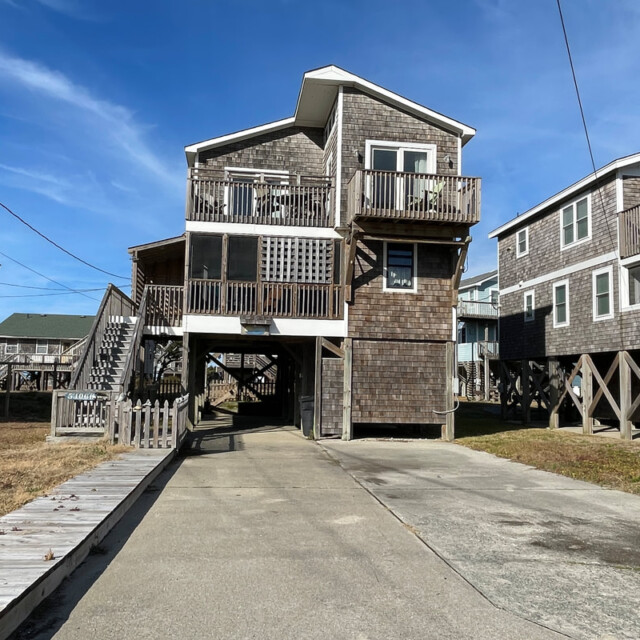 A two-story, wood-paneled beach house on stilts with balconies, driveway, and blue sky in the background.