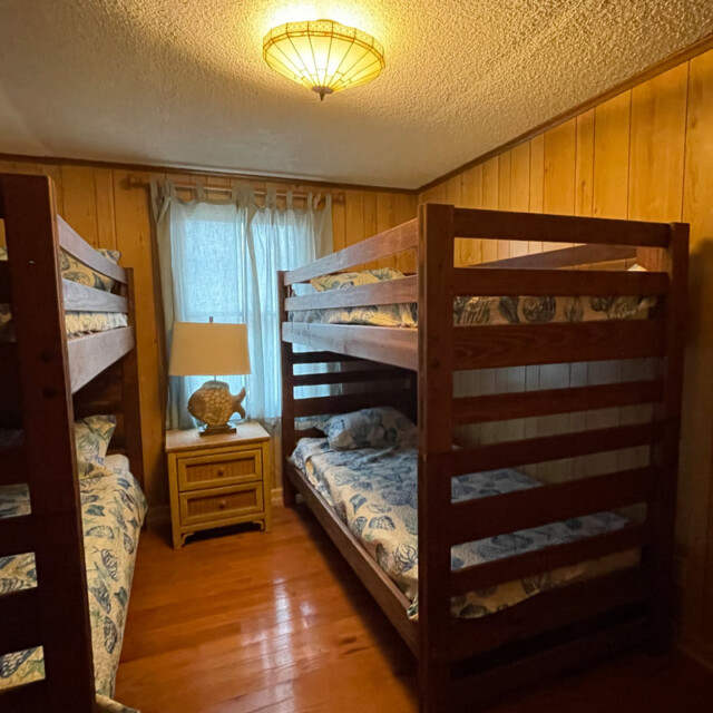 Wood-paneled room with two wooden bunk beds, blue-patterned bedding, window, lamp, and small nightstand.