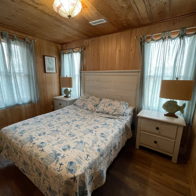 Wood-paneled bedroom with a bed, two nightstands, fish-shaped lamps, and sheer curtains on the windows.
