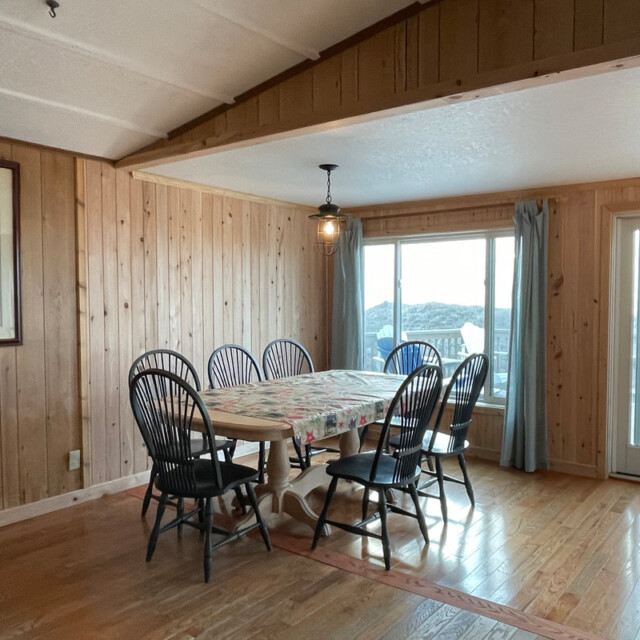 Dining area with a wooden table, six chairs, wood-paneled walls, large windows, and a framed bird picture.