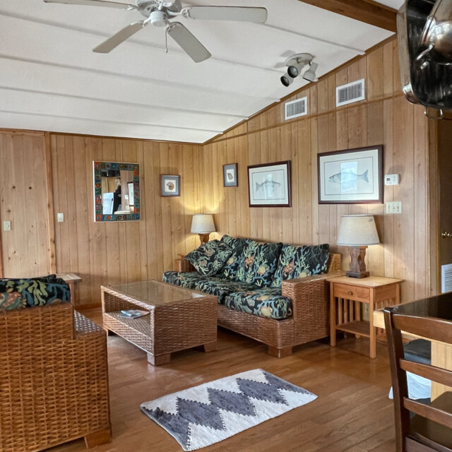 Wood-paneled living room with wicker furniture, floral upholstery, a ceiling fan, lamps, and framed art on the walls.