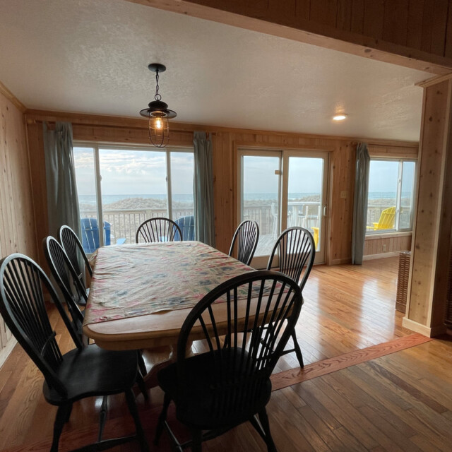 Dining table with six chairs in a wood-paneled room, large windows, and ocean view in the background.
