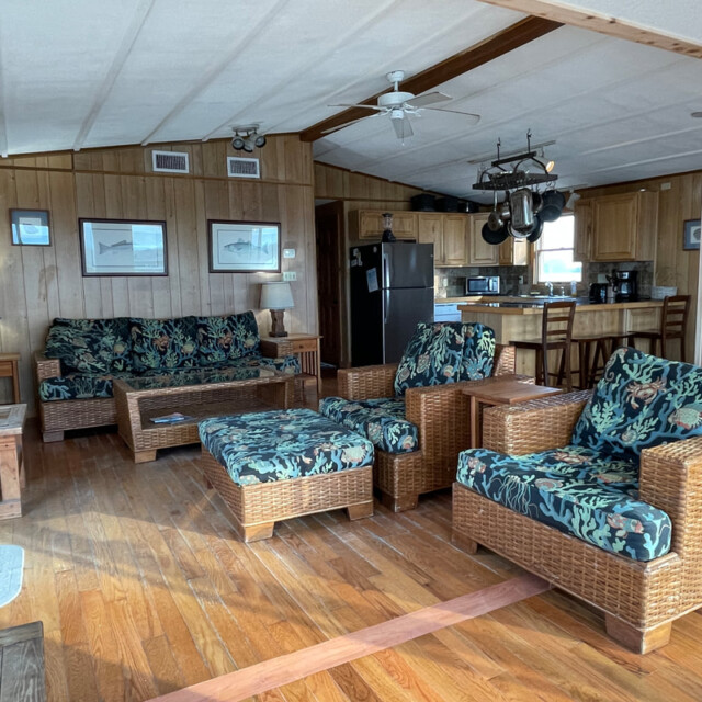 Living room with wicker furniture, floral cushions, hardwood floors, and a view of the kitchen in a wood-paneled house.