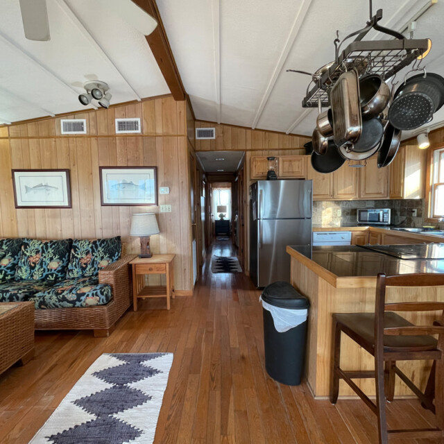 Open kitchen and living area with wood paneling, wicker furniture, hanging pots, and stainless steel appliances.