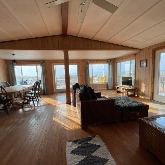 Sunlit living and dining area with wood walls, large windows, and ocean views, furnished with wicker and wood furniture.