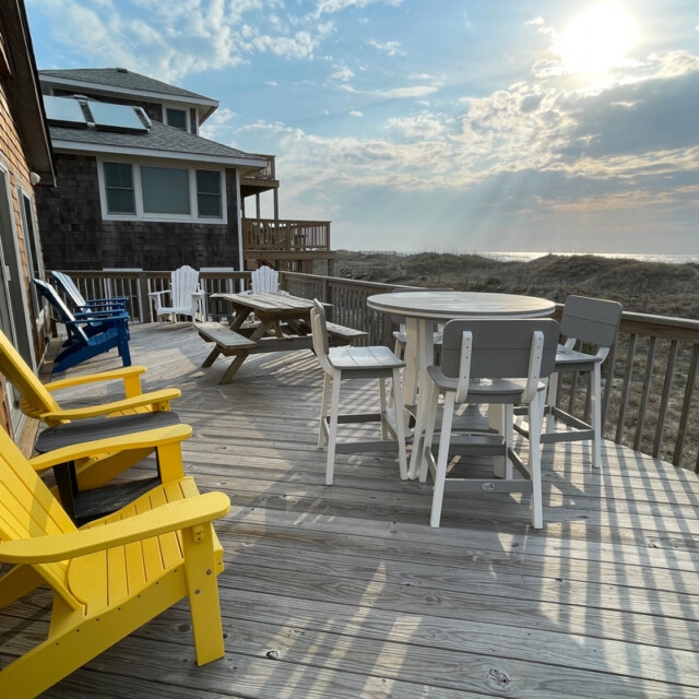 Wooden deck with yellow and white chairs, a table, and ocean view under a partly cloudy sky at sunrise or sunset.