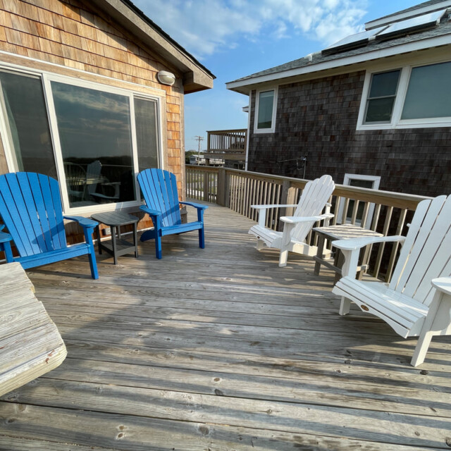 Wooden deck with two blue and two white Adirondack chairs, a small table, and two shingle-sided houses.