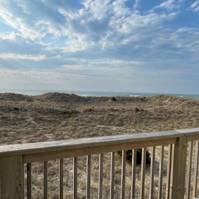Wooden deck with railing overlooks sandy dunes, sparse grass, and ocean under a partly cloudy blue sky.